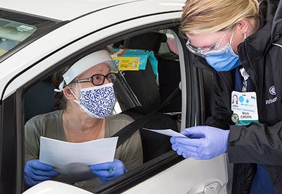 Masked woman sits in car driver's seat at drive through event, speaking with health worker outside car