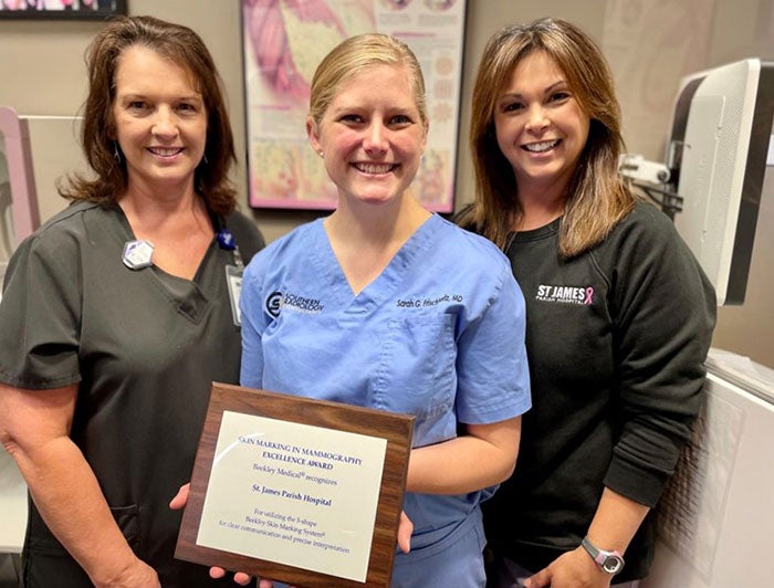 Three women posing for a picture, smiling, and wearing scrubs while the woman in the middle is holding a plaque award 