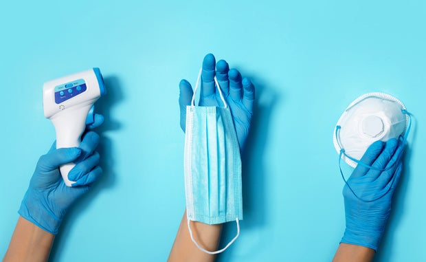 4 Crisis Management Steps to Reduce Supply Disruption. Three hands wearing blue nitrile surgical gloves hold a digital thermometer, a surgical mask, and an N95 respirator.